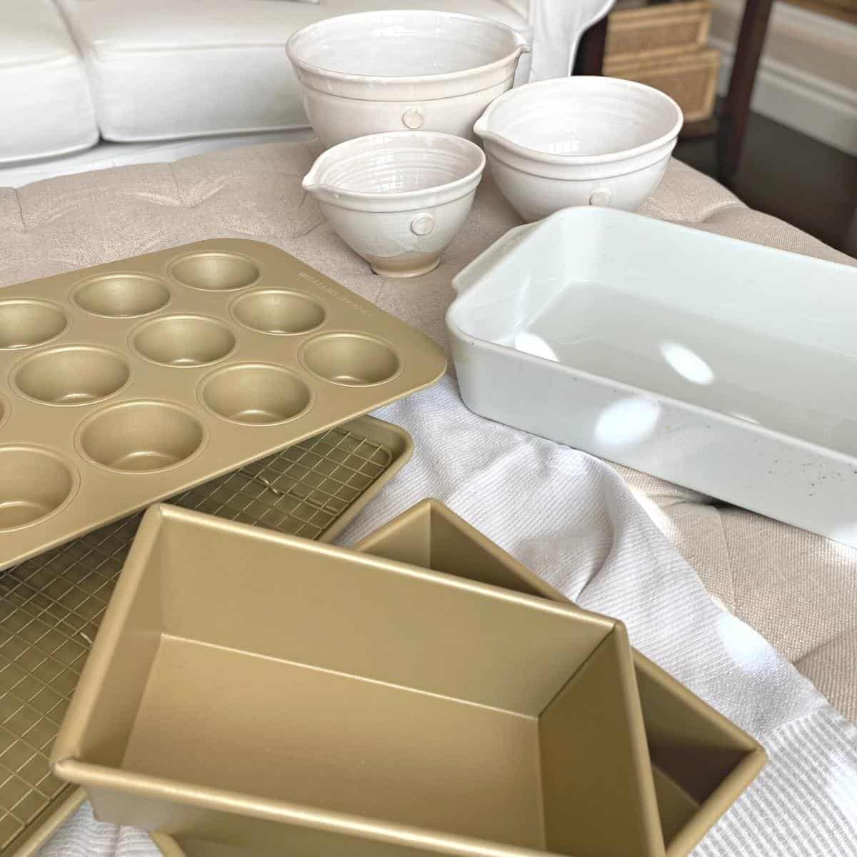 Gold bakeware set, white pottery mixing bowl set and a white roaster on a neutral kitchen towel