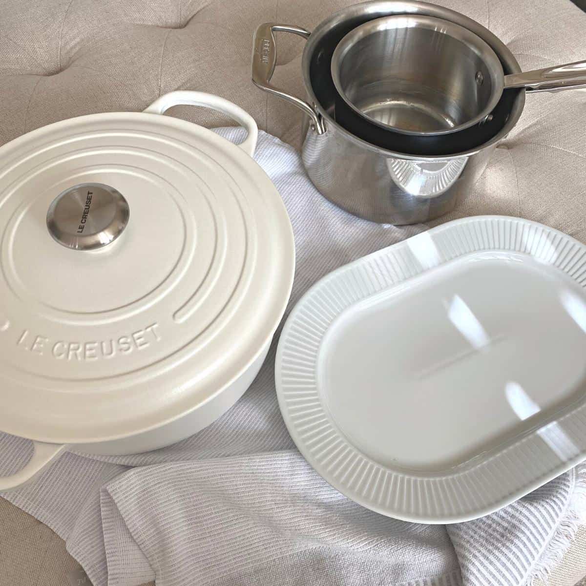 White cast iron pot, white serving platter and stainless steel pots on a neutral cotton towel