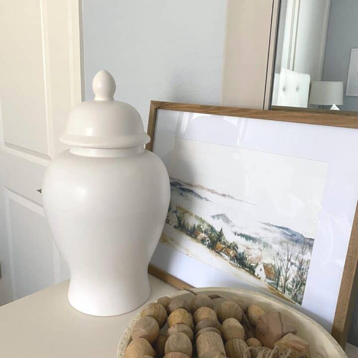 Large white ginger jar on a dresser in front of a mountainside artwork and bowl with wood bead decor.