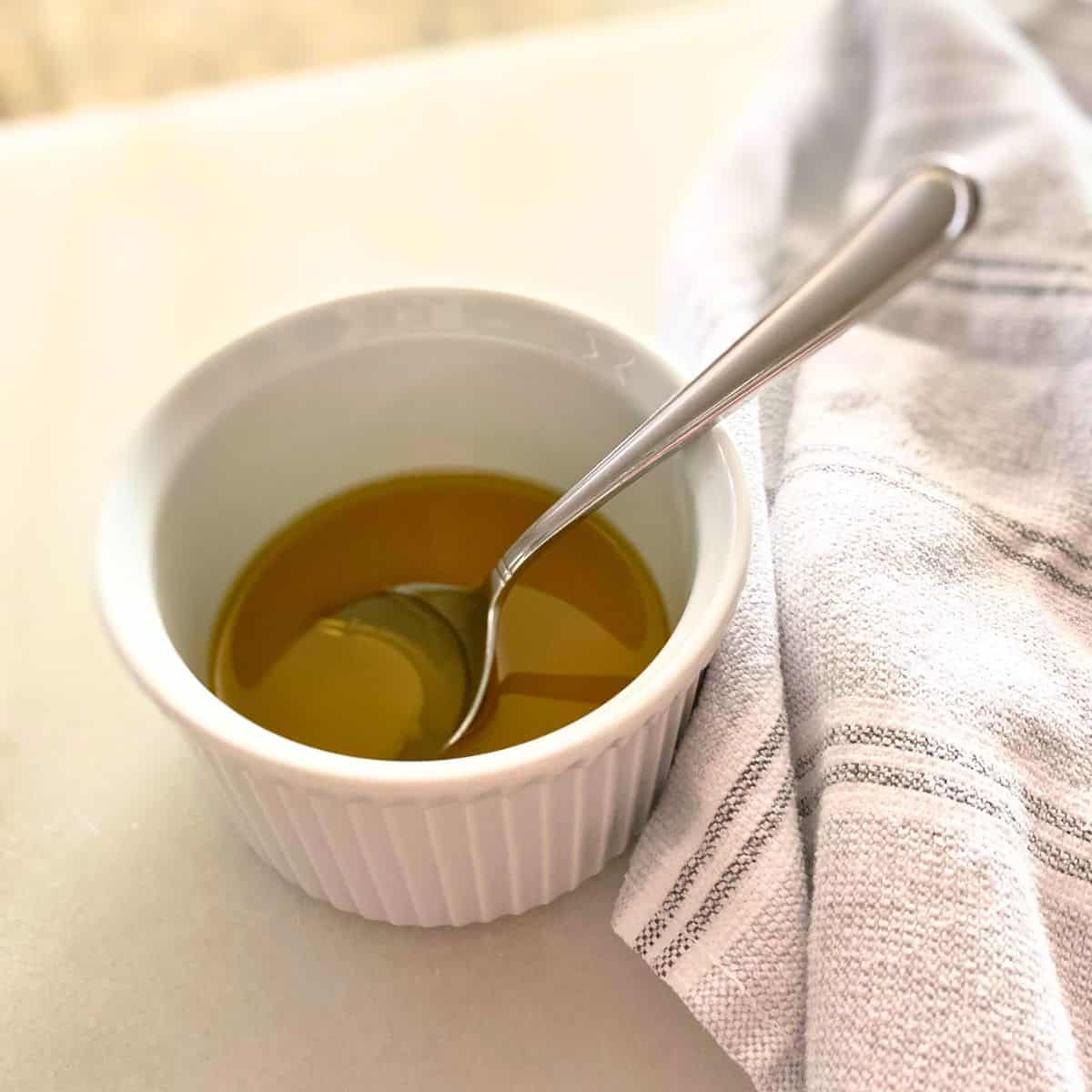 Lemon olive oil dressing in a white ramekin with a stainless steel soup spoon and a neutral cotton kitchen towel next to it