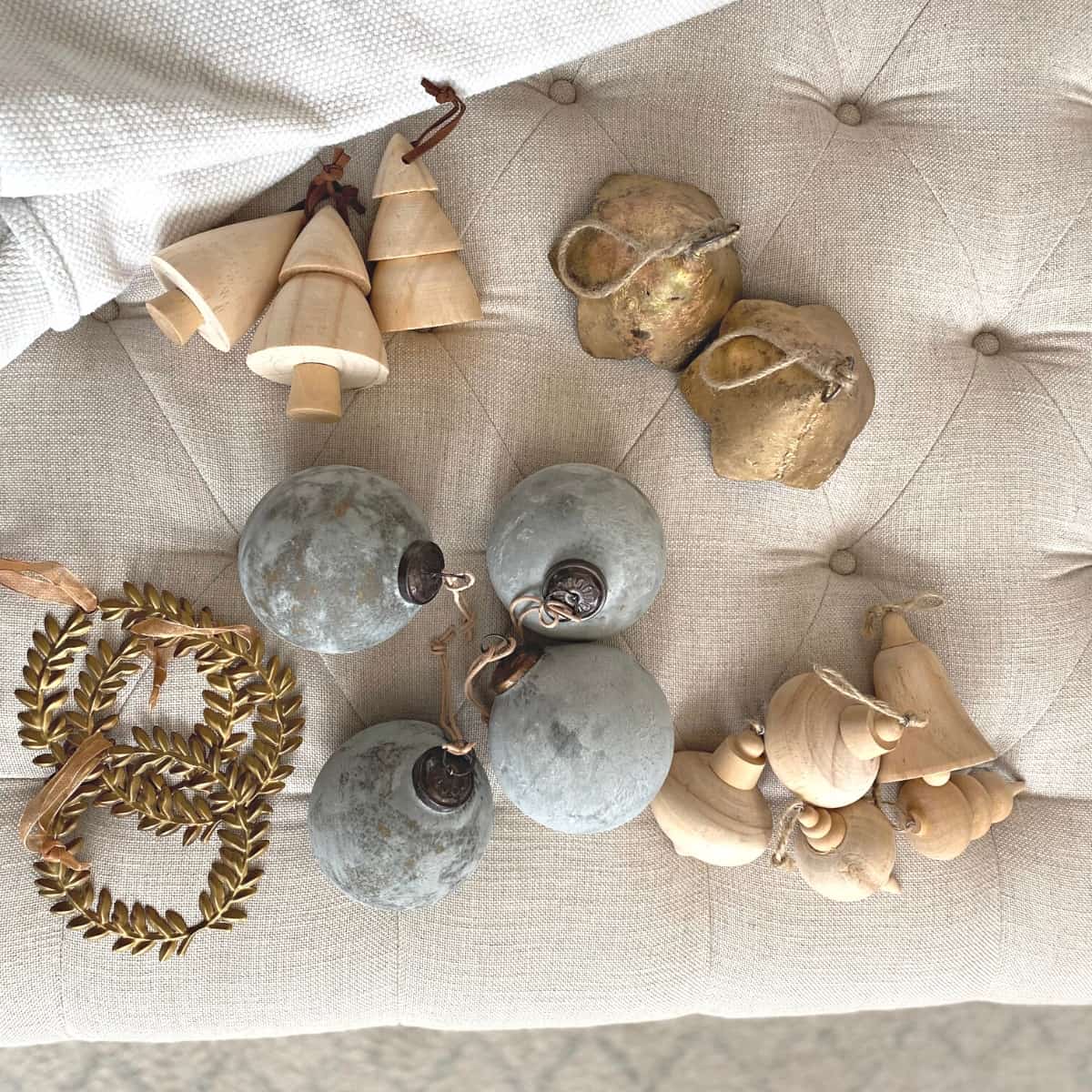 Neutral Christmas ornaments of brass, wood and dusty blue atop a linen tufted ottoman