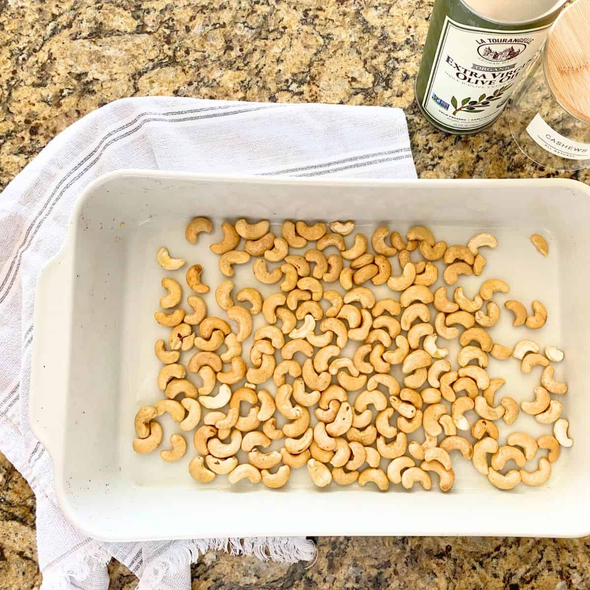 Raw cashews in a white roasting pan next to olive oil and an empty glass jar with a white label and the word cashews