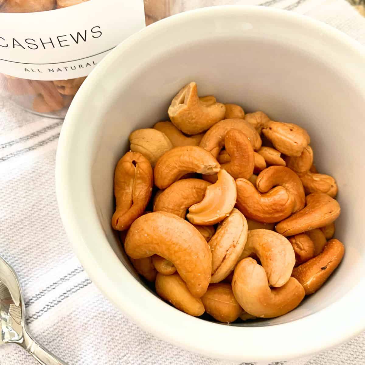 Roasted cashews served in a white ramekin next to a glass jar with a white label and the word cashews