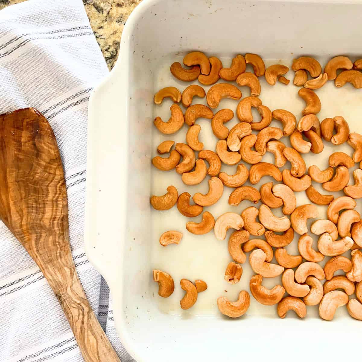Roasted cashews in a white porcelain roasting pan, with a wooden spatula resting beside on a light colored kitchen towel