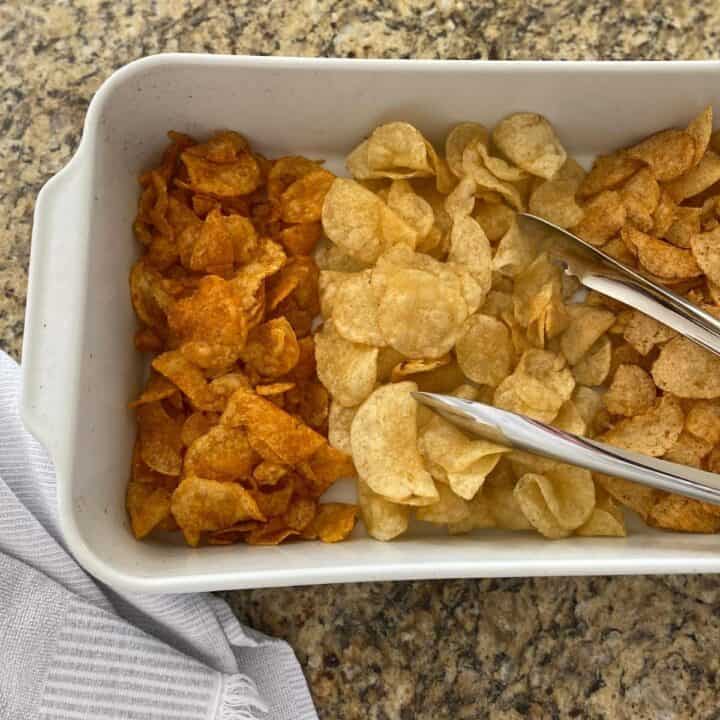 Three different flavors of potato chips served in a white rectangular roasting pan with stainless steel serving tongs