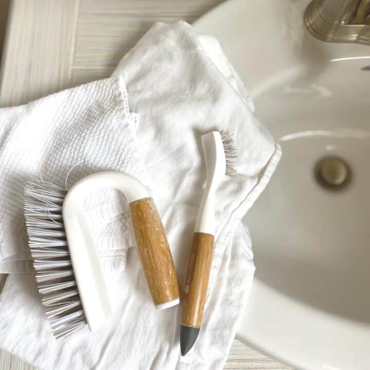 White, wood-handled scrub brush and grout brush for cleaning, sitting on top of white cotton cleaning cloths.