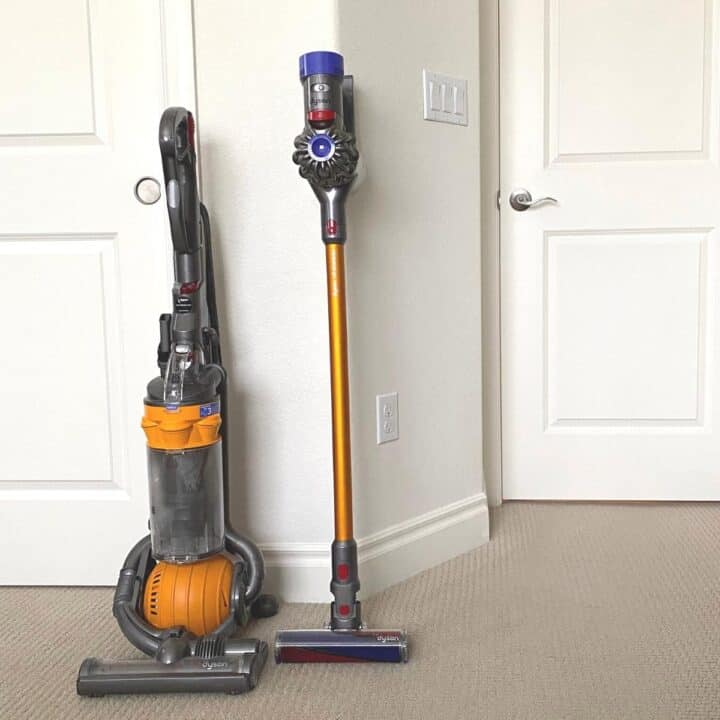 Two Dyson vacuum cleaners propped against a white bedroom wall. One is an upright vacuum, while the other is a stick vacuum.