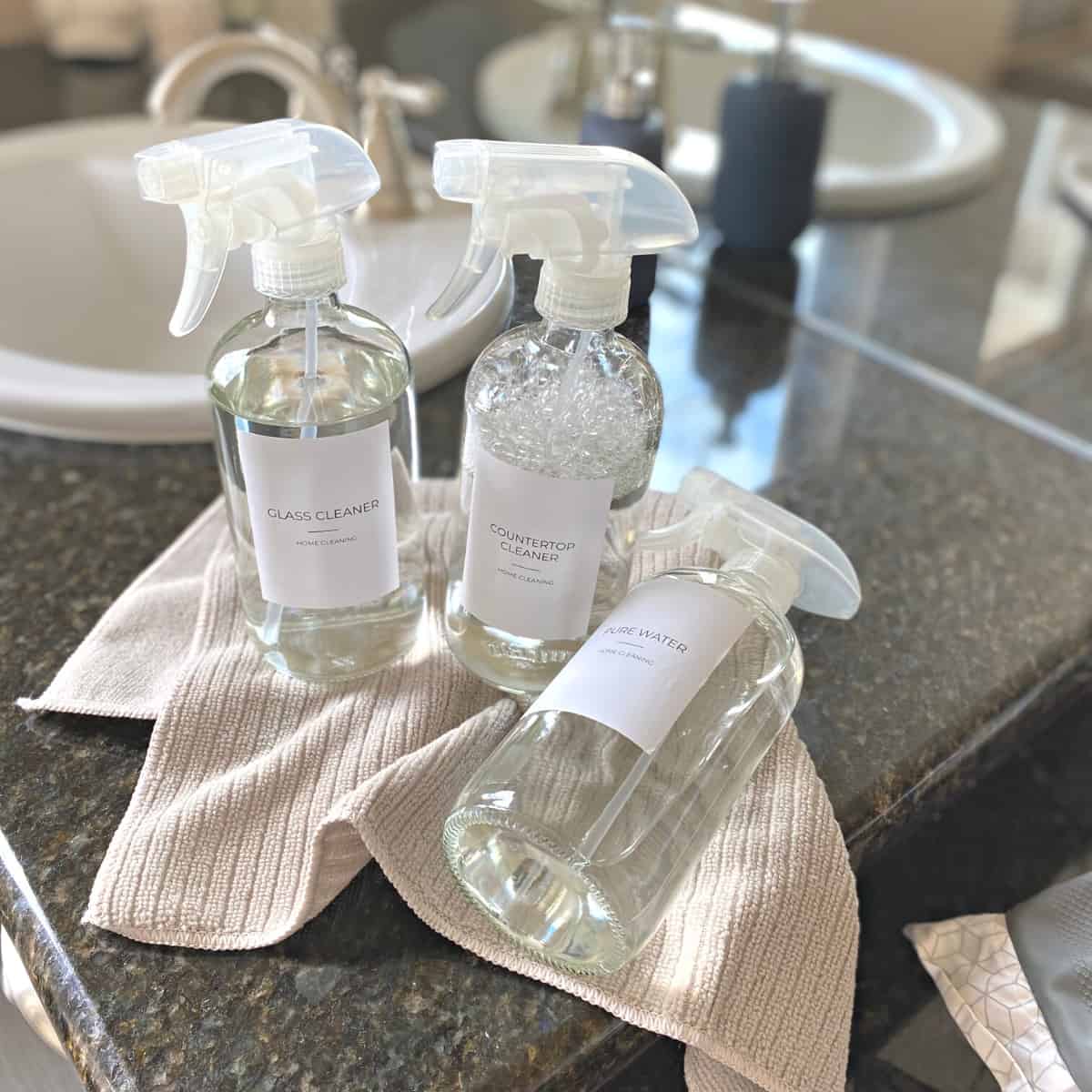 Glass spray bottles of bathroom cleaning products lay on a dark granite countertop